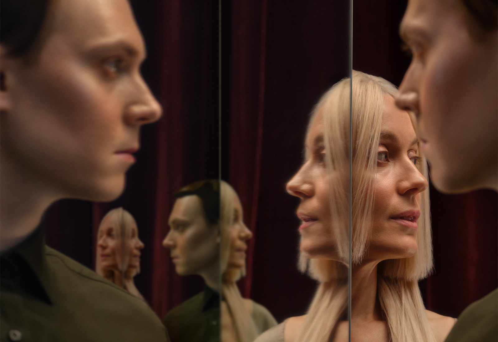 Kaksi ihmistä, toinen vaaleatukkainen ja toinen tummahiuksinen, seisovat vastakkain peilien edessä, jolloin heidän kasvonsa heijastuvat useaan kertaan tummanpunaista verhotaustaa vasten.