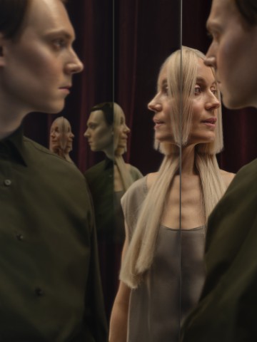 Mies ja nainen seisovat peilipaneelien vieressä, jolloin heidän kasvonsa ja ylävartalonsa heijastuvat useaan kertaan. Taustalla on tummat viininpunaiset verhot, ja molemmat kuvattavat näyttävät mietteliäiltä.