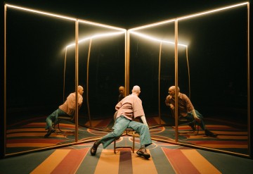 Henkilö satunnaisissa vaatteissa istuu tuolilla, nojautuu taaksepäin jalat levällään, ja häntä ympäröivät suuret peilit, jotka luovat useita heijastuksia. Lämpimät ylävalot ja värikäs raidallinen lattia lisäävät dramaattista tunnelmaa.