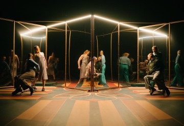 Useat ihmiset ovat vuorovaikutuksessa ja poseeraavat geometrisen peilirakenteen ympärillä, jossa on lämpimät valot, jotka luovat useita heijastuksia. Kohtaus on asetettu värikkäälle kuviolliselle lattialle, mikä antaa surrealistisen, taiteellisen ilmapiirin.