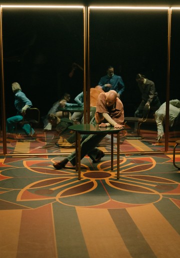 Kalju henkilö oranssissa paidassa nojaa pöydän yli kuviollisella lattialla, ja taustalla on useita ihmisiä peilattuna, mikä luo dramaattisen, teatraalisen kohtauksen.