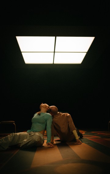 Kaksi ihmistä istuu värikkäällä lattialla kirkkaan neliön muotoisen kattovalaisimen alla pimeässä huoneessa. Toinen, siniseen toppiin pukeutunut, nojaa taaksepäin, kun taas toinen, beigeen pukeutunut, nojaa heitä kohti, kasvot lähellä toisiaan.