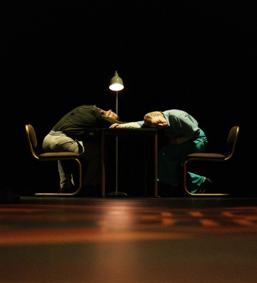 Kaksi ihmistä istuu toisiaan vastapäätä pöydän ääressä, molemmat eteenpäin lyyhistyneinä ja päät pöydällä yhden lampun alla, mikä luo dramaattisen ja tunnelmallisen tunnelman pimeään huoneeseen.