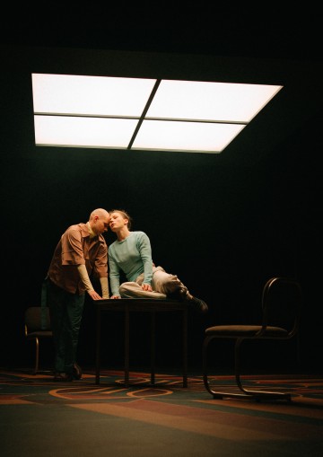 Kaksi ihmistä on vuorovaikutuksessa suuren neliönmuotoisen kattovalaisimen alla; toinen istuu pöydällä ja toinen nojaa lähelle. Kaksi tyhjää tuolia on lähellä hämärässä huoneessa, jossa on kuviollinen lattia.