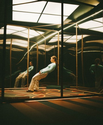 Vaaleisiin housuihin ja vaaleansiniseen villapaitaan pukeutunut nainen nojaa taaksepäin peilihuoneessa, jossa on kultakehykset ja geometriset kattovalaisimet, jotka luovat useita heijastuksia hänen ympärilleen.