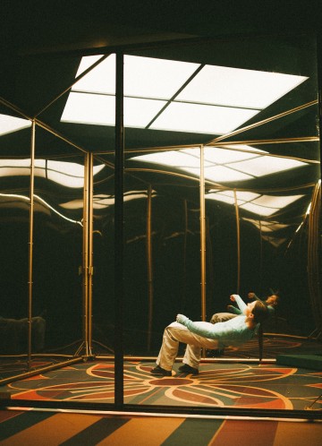 Rento vaatteisiin pukeutunut henkilö makaa tuolilla huoneessa, jonka peiliseinät, kuviollinen lattia ja suuret hehkuvat kattovalaisimet luovat surrealistisen, heijastavan ilmapiirin.