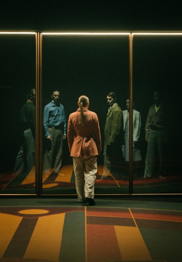 Poninhäntäinen henkilö, jolla on punainen takki ja vaaleat housut, seisoo heijastavaa lasiseinää vasten, jossa näkyy hänen ja kolmen muun, erilaisiin liikeasuihin pukeutuneen henkilön heijastukset.