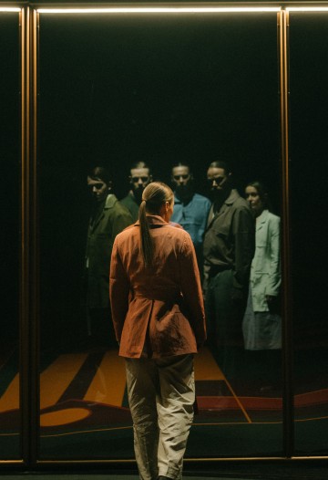 Henkilö, jolla on poninhäntä, seisoo lasiseinää vasten, jossa heijastuu neljä tummiin vaatteisiin pukeutunutta hahmoa, jotka seisovat yhdessä hämärässä, dramaattisessa ympäristössä.