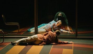 Siniseen asuun pukeutunut henkilö makaa lattialla ja nojaa peiliin, jossa on hänen peilikuvansa ja toinen hahmo taustalla. Toinen vaaleanpunaiseen takkiin pukeutunut henkilö makaa niin ikään kuvioidulla lattialla lähellä.