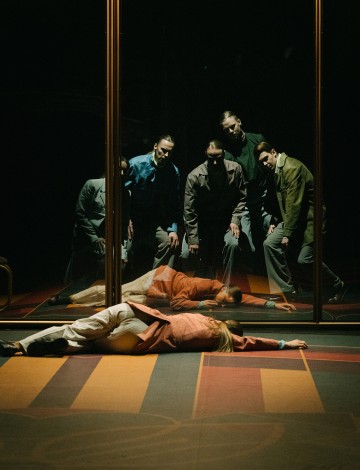 Viisi ihmistä seisoo peilin edessä ja katsoo kahta muuta lattialla makaavaa ihmistä. Heidän heijastuksensa ovat näkyvissä, ja kohtaus on hämärästi valaistu, ja siinä on dramaattisia varjoja ja värillisiä lattiakuvioita.