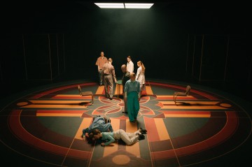 Kahdeksan ihmistä seisoo kuvioidulla lattialla kirkkaiden valojen alla, ja etualalla kaksi makaa lähekkäin. Heidän ympärillään on neljä tuolia, ja kohtauksessa on dramaattinen, teatraalinen tunnelma.