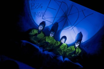 Neljä kirkkaankeltaisiin pukuihin pukeutunutta ihmistä istuu vierekkäin sinisen valon valaisemina. Heidän kasvonsa ovat ilmeikkäät, ja heidän takanaan olevaa seinää peittää graffitin kaltainen kirjoitus. Kohtaus vaikuttaa teatraaliselta ja surrealistiselta.