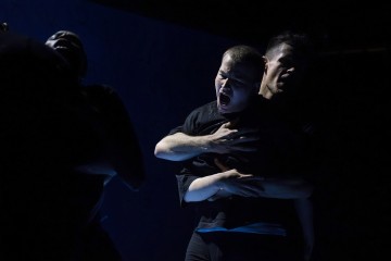 Dramaattisessa valaistuksessa oleva henkilö puristaa rintaansa ja huutaa, kun toinen hahmo seisoo tiiviisti hänen takanaan ja osittain syleilee tai hillitsee häntä. Tausta on tumma, mikä korostaa tunteiden voimakkuutta.