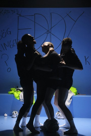 Neljä tummiin vaatteisiin pukeutunutta ihmistä seisoo tiiviisti yhdessä lavalla, jossa on sininen valaistus, ja heidän kasvonsa ovat varjossa. Heidän takanaan olevalla seinällä näkyy graffitin kaltaista kirjoitusta, ja vaatteet ovat hajallaan lattialla.