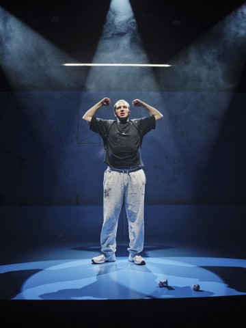 Henkilö seisoo lavalla valonheittimen alla ja taivuttaa molempia käsiään. Hänellä on yllään musta paita, vaaleanharmaat verkkarit ja lenkkarit. Taustalla on sininen graffitimainen kirjoitus ja vaaleaa savua. Lattialla on kaksi rypistynyttä paperia.