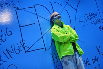 Kirkkaaseen neonväriseen takkiin pukeutunut henkilö seisoo hymyillen ja kädet ristissä nojaamassa siniseen seinään, joka on peitetty suurilla, mustilla graffitityylisillä kirjoituksilla ja doodleilla.