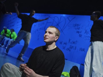 Mustapaitainen henkilö istuu lattialla ja katsoo ylöspäin, kun taas muut seisovat tai liikkuvat sumeassa sinisellä valaistussa taustassa. Graffitityylinen kirjoitus peittää heidän takanaan olevan sinisen seinän.