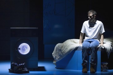 Valkoiseen t-paitaan ja farkkuihin pukeutunut nuori mies istuu sängyn reunalla hämärässä huoneessa, vastapäätä pesukonetta, jonka ovi on valaistu. Sängyn alle on pinottu kirjoja, ja hänen takanaan olevaa seinää peittää graffiti.