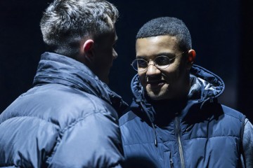 Kaksi tummiin takkeihin pukeutunutta nuorta miestä kohtaa toisensa hämärässä. Toinen hymyilee ja käyttää pyöreitä silmälaseja, kun taas toinen on selkä kameraan päin. Tunnelma vaikuttaa vakavalta tai intensiiviseltä.