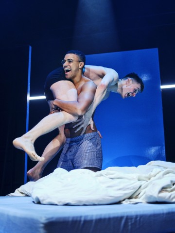 Kaksi alusvaatteisiin pukeutunutta miestä nauraa ja leikkii sängyllä, ja toinen mies nostaa toista sylissään. Tausta on dramaattisesti valaistu sinisen ja valkoisen sävyillä.