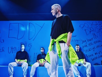 Lavalla on neljä ihmistä, joilla on samanlaiset mustat paidat ja harmaat verkkarit, yksi seisoo edessä ja kolme istuu takana. Heidän takanaan olevaa sinistä seinää peittää lihavoitu graffitityylinen teksti. Valaistus on dramaattinen ja teatraalinen.