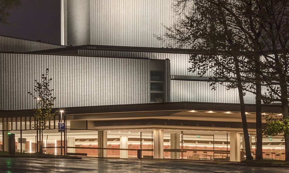 Moderni rakennus, jonka lasiseinät on valaistu yöllä, jossa on selkeät linjat ja näkyvä uloke sisäänkäynnin yläpuolella, etualalla muutama puu ja märkä jalkakäytävä.