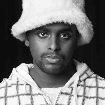 Mustavalkoinen muotokuva henkilöstä, jolla on pörröinen pipo ja ruudullinen huppari ja joka katsoo suoraan kameraan neutraalilla ilmeellä. Tausta on tumma.