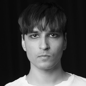 Nuori mies, jolla on suorat tummat hiukset ja vakava ilme, katsoo suoraan kameraan. Hänellä on vaalea paita, ja tausta on tumma. Kuva on mustavalkoinen.