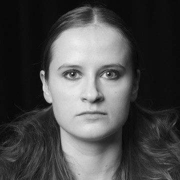 Mustavalkoinen muotokuva nuoresta naisesta, jolla on suorat, keskeltä erotetut hiukset ja joka katsoo suoraan kameraan neutraalilla ilmeellä tummaa taustaa vasten.