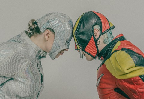 Kaksi futuristisiin asuihin pukeutunutta ihmistä, joista toisella on kiiltävä hopeinen asu ja kypärä ja toisella punainen, keltainen ja musta kypärä ja puku, seisovat vastakkain otsat vastakkain.