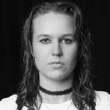 Henkilö, jolla on märät, olkapäiden pituiset hiukset ja neutraali ilme, on kameraan päin. Hänellä on yllään choker-kaulakoru ja valkoinen toppi. Tausta on tumma. Kuva on mustavalkoinen.