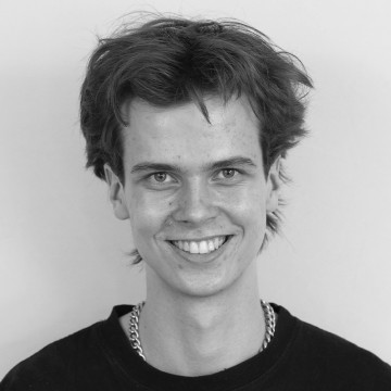 Nuori henkilö, jolla on lyhyet, sekaiset hiukset, hymyilee kameralle. Hänellä on yllään tumma paita ja ketjukoru. Tausta on tavallinen ja vaalea. Kuva on mustavalkoinen.