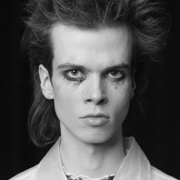 Nuori henkilö, jolla on sekaiset hiukset ja dramaattinen, mustaksi tahriintunut silmämeikki, tuijottaa suoraan kameraan. Hänellä on vaalea, kauluspaita, ja tausta on tumma.