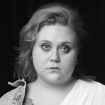 Henkilö, jolla on vaalea iho ja aaltoilevat, olkapäiden pituiset hiukset, katsoo suoraan kameraan neutraalilla ilmeellä. Hänellä on korvakorut ja vaalea toppi tummaa taustaa vasten.