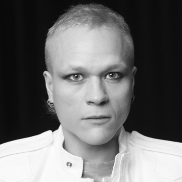 Henkilö, jolla on lyhyet vaaleat hiukset, valkoinen takki ja pienet korvakorut, katsoo suoraan kameraan neutraalilla ilmeellä mustaa taustaa vasten. Kuva on mustavalkoinen.