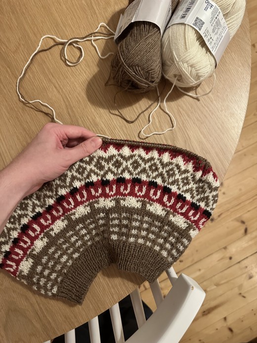 Käsi pitelee pyöreää, osittain neulottua, ruskeaa, valkoista, punaista ja mustaa lankaa olevaa kuviopuseroa. Puupöydällä lepää kaksi lankapalloa, joista toinen on ruskeaa ja toinen kermanväristä. Valkoinen tuoli istuu lähellä puulattialla.