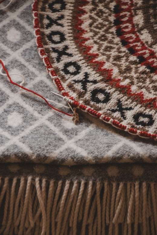 Lähikuva kuvioidusta kudotusta matosta, jossa on punaisia, mustia ja ruskeita kuvioita ja joka on osittain viimeistelty punaisilla ja valkoisilla langoilla. Matto lepää harmaan ja valkoisen hapsutetun tekstiilin päällä.