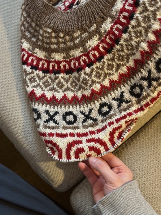 Käsi pitää kiinni neulepuseron reunasta, jossa on monimutkaisia kuvioita ruskealla, kermanvärisellä, punaisella ja mustalla. Puserossa on geometrisia ja XOXO-kuvioita, ja se lepää kuvioidun beigen kankaan päällä.