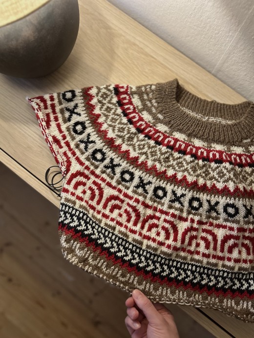 Kädessä on värikäs neulepusero, jossa on monimutkaisia punaisia, mustia ja valkoisia kuvioita, muun muassa X- ja O-kuvioita, ja joka on vaalealla puupöydällä pyöreän harmaan lampun vieressä.