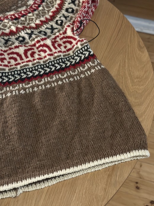 Lähikuva puupöydällä olevasta osittain neulotusta villapaidasta. Vaatteessa on punaisen, mustan ja kermanvärisiä monimutkaisia kuvioita yläosassa ja yksivärinen ruskea alaosa, jonka alareunassa on kermanvärinen reuna.