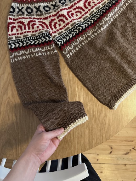 Käsi pitelee ruskeaa neulepuseroa, jossa on kermanvärisiä, punaisia ja mustia kuvioita rinnassa ja hihassa. Se on esillä pyöreällä puupöydällä valkoisen tuolin vieressä.