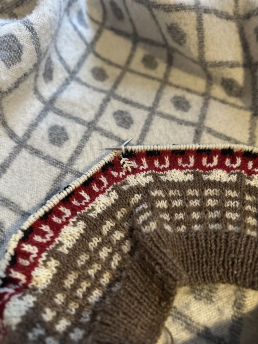 Lähikuva neuleprojektista, jossa on ruskeita, kermanvärisiä, punaisia ja valkoisia kuvioita ja joka lepää harmaan ja valkoisen peiton päällä, jossa on geometrinen kuvio. Neulepuikot on työnnetty silmukoihin projektin yläosassa.