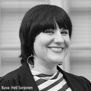 Lyhyet tummat hiukset omaava nainen hymyilee, yllään raidallinen paita, tumma takki ja suuret korvakorut. Tausta on sumea. Alareunassa on teksti: Kuva: Heli Sorjonen.