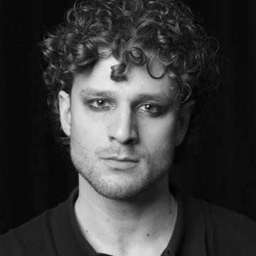 Mustavalkoinen muotokuva henkilöstä, jolla on kiharat hiukset ja hienovarainen silmämeikki, päällään tumma paita, katselee suoraan kameraan neutraali ilme tummaa taustaa vasten.