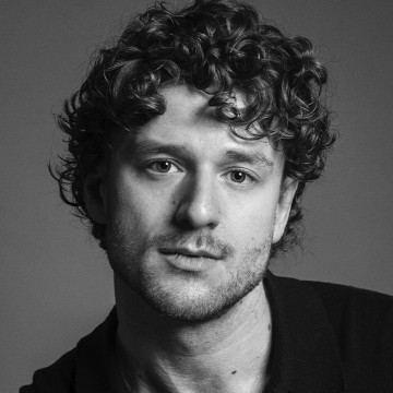 Mustavalkoinen muotokuva nuoresta miehestä, jolla on lyhyet, kiharat hiukset ja vaaleat karvat, ja joka katsoo suoraan kameraan neutraalilla ilmeellä. Hänellä on yllään tumma kauluspaita tavallista taustaa vasten.