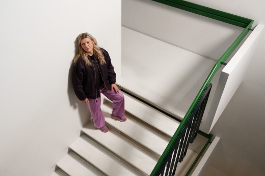 Pitkä vaaleatukkainen henkilö, jolla on musta takki ja violetit housut, seisoo valkoisella portaikolla seinän vieressä ja katsoo ylöspäin. Portaiden kaiteet ovat mustat ja vihreät. Ympäristö vaikuttaa vähäeleiseltä ja modernilta.