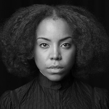 Mustavalkoinen muotokuva nuoresta naisesta, jolla on luonnolliset, tuuheat ja kiharat hiukset, yllään korkeakauluksinen, kuvioitu tumma toppi, katselee suoraan kameraan neutraalilla ilmeellä tummaa taustaa vasten.