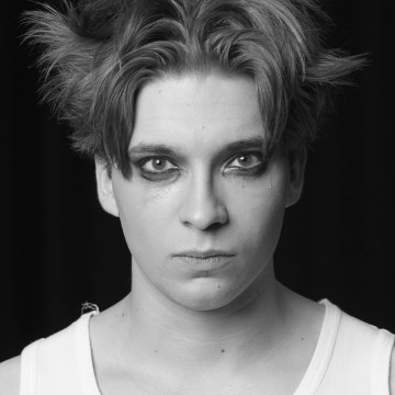 Mustavalkoinen valokuva henkilöstä, jolla on sekaiset hiukset, tumma silmämeikki ja vakava ilme, yllään hihaton valkoinen paita tummaa taustaa vasten.