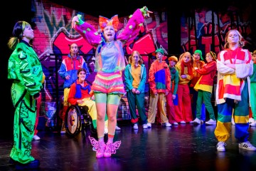 Ryhmä värikkäisiin, eklektisiin pukuihin pukeutuneita esiintyjiä seisoo lavalla, jonka taustalla on graffitilla päällystetty tiiliseinä. Keskeinen esiintyjä, jolla on päällään kirkkaan vaaleanpunainen ja vihreä, seisoo kädet kohotettuna, kun muut katselevat ja reagoivat.