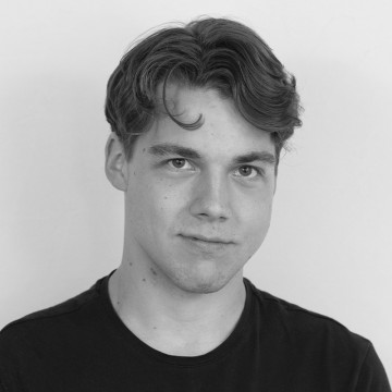 Nuori mies, jolla on lyhyet, aaltoilevat hiukset ja tumma t-paita, katsoo kameraan neutraalilla ilmeellä. Kuva on mustavalkoinen ja taustaltaan tavallinen.